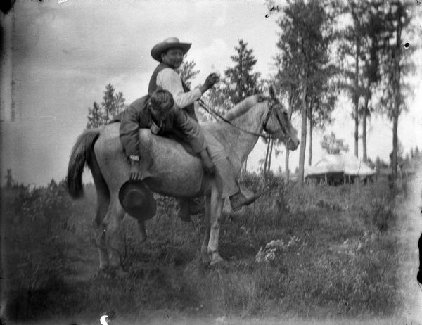 Ho-Chunk man wearing hat mounted on horse. He is holding what appears to be a glass bottle in his right hand. Another Ho-Chunk man is lying face down across the rear of the horse, holding his hat. Identified as Luke Green posing sitting, and David Good Village hanging across the back. In the background is an awning on stakes among trees.