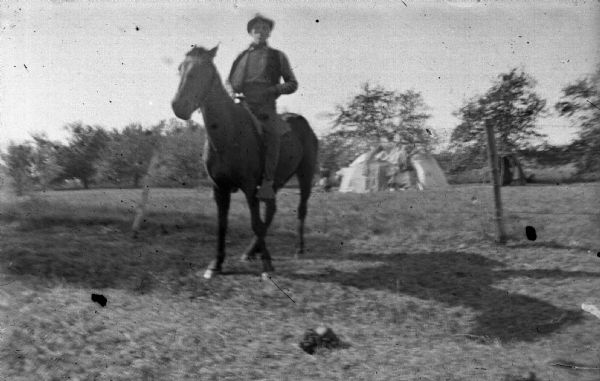 Ho-Chunk man sitting on a horse in a field. In the background near trees is a lodge.