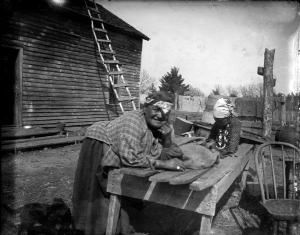 Two Ho-Chunk women are leaning on a rough-hewn table on the ground in front of a wooden building. Against the building is a ladder leading to the roof. The woman on the left is wearing a headache band and plaid shirt and is identified as probably the first wife of Joe Bear Heart. A dog is standing near her on the left.