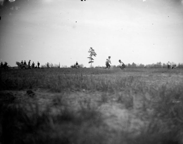 View across field towards men running and standing in a field in the distance, probably a baseball game. There are people sitting in horse-drawn wagons on the right, perhaps watching the game.