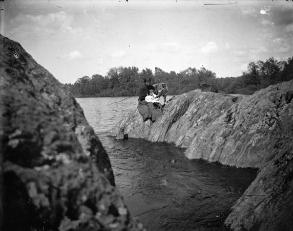 View across rocks along a river towards a Ho-Chunk woman, child, and dog posing on the large, steep rocks on the shoreline.