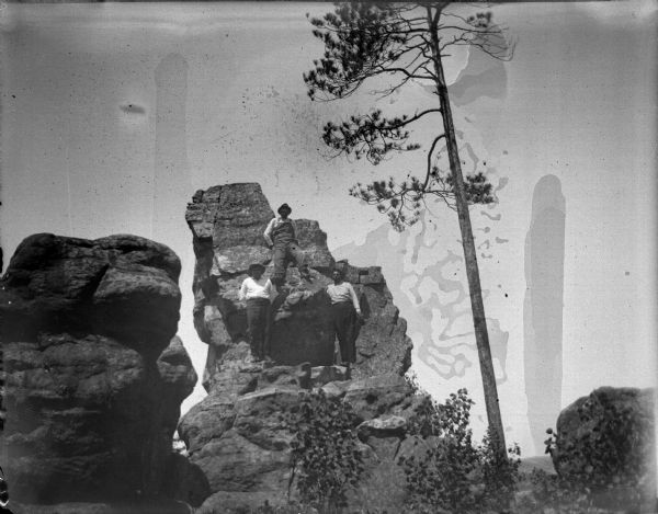 View looking up towards three Ho-Chunk men posing standing on a rock face near the top of a rock pillar near a tall tree. Two men are wearing hats and the other is holding his hat in his left hand.