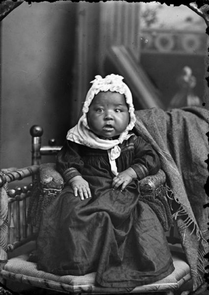 Studio portrait of a Ho-Chunk infant girl posing sitting in a small chair that has been placed on top of another chair. She is in front of a painted backdrop and is wearing a dark dress, light-colored bandana, and bonnet.