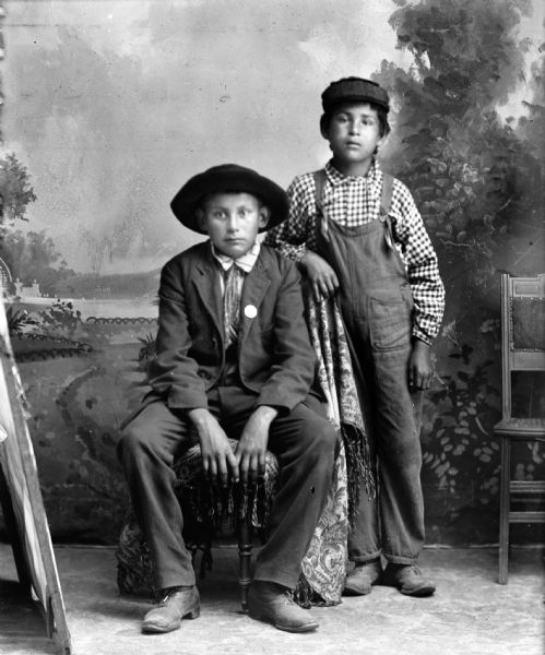 Studio portrait in front of a painted backdrop of a Ho-Chunk boy posing sitting in a chair on the left wearing a suit coat and hat. Another Ho-Chunk boy is posing standing beside him on the right, with his right arm resting on the back of the chair. He is wearing bib overalls, checked shirt, and a cap.