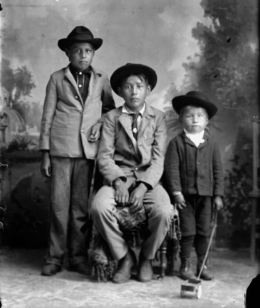 Studio portrait of three Ho-Chunk boys posing in front of a painted backdrop. In the center a Ho-Chunk boy is sitting wearing a suit, hat, and necktie. Another Ho-Chunk boy is standing on the left wearing a suit coat and hat, and a small Ho-Chunk boy on the right is wearing a winter coat and hat, and holding the handle of a toy wagon.