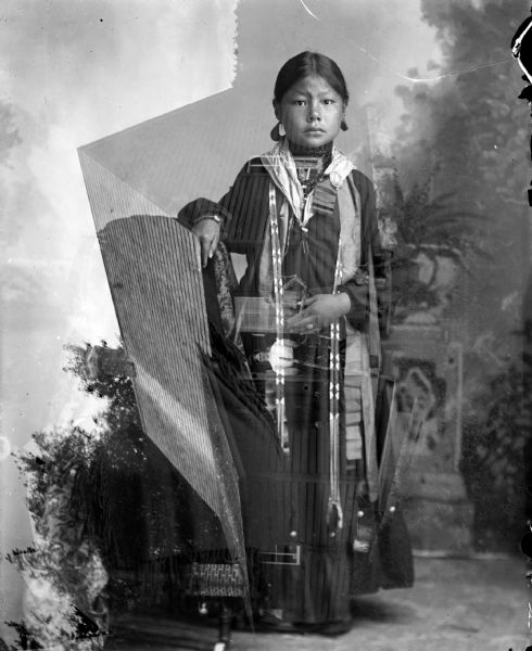 Double exposure, one exposure of a studio full-length portrait of a Ho-Chunk girl posing standing in front of a painted backdrop wearing several necklaces, earring, and streamers. The other exposure is a horizontal image of a woman posing standing in the doorway of a wooden house.  	
