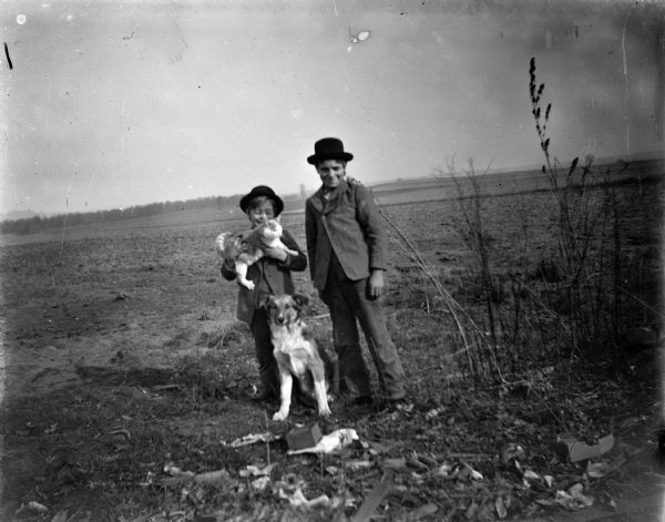 Two European American or Ho-Chunk boys wearing hats are posing standing in a field with a dog and cat. On the far horizon are trees and low hills.