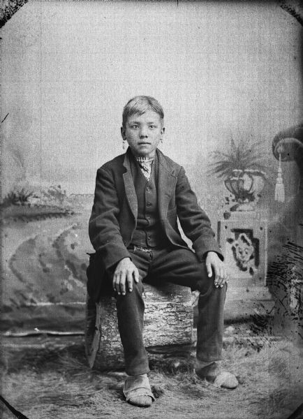 Studio portrait of George Bearchief (Ha Na Kah) posing seated on a hollow log in front of a painted backdrop. He has his hands on his knees and is wearing a suit coat, vest, trousers, moccasins, and earrings.