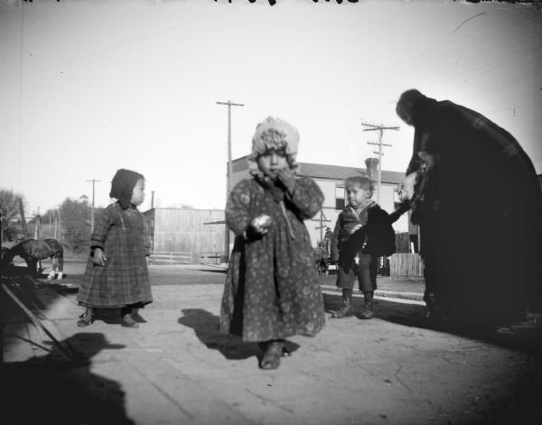 Ho-Chunk woman, and three Ho-Chunk children wearing winter clothes, standing on a board sidewalk in town, probably Black River Falls. Two of the children are wearing dresses and bonnets. In the background are buildings, power lines, and a horse.