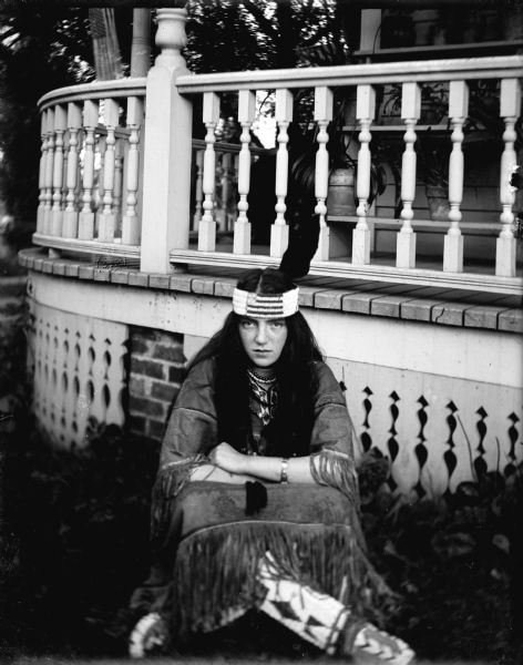 A European American woman dressed in Ho-Chunk regalia, including a fringed dress, necklaces, and headband with a feather, posed sitting cross-legged on the ground in front of a veranda. Identified as Capitola Maddock in front of the veranda of Will Maddock.