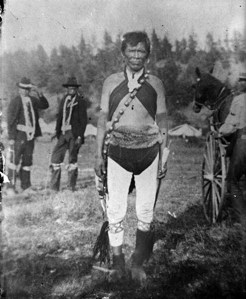 A Ho-Chunk man is posing standing in a field, holding an object in his left hand. He is wearing regalia including a long bandoleer which appears to have bells attached, light-colored leggings, a fur belt, and dark breech cloth. He is identified as George Standing Water of the Blue Wing Band, clan name Water Spirit Little Ocean Waves, circa 1907. In the background there are two men standing on the left, and a horse pulling a wagon or carriage on the right. Tents are in the far background near a hill with trees.