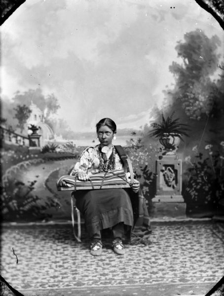 Studio portrait of a European American girl posing sitting in front of a painted backdrop, holding a Ho-Chunk cradle board with a china-headed doll. She is wearing Ho-Chunk necklaces, gorget, moccasins, and shawl. Identified as Julia Maddock wearing items from the collection of Tom Roddy.