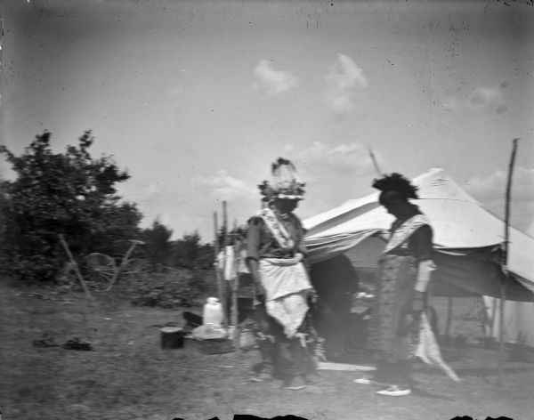 Two Ho-Chunk men dressed in a regalia standing in front of a tent, possibly at a powwow.