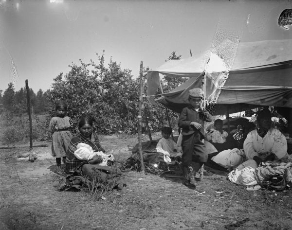 Group of people under and near a tent. One Ho-Chunk woman is sitting outside a canvas awning holding an infant, with a Ho-Chunk girl standing behind her. A young boy is standing in the middle wearing a cap, jacket, and short trousers. Another Ho-Chunk woman and several children are also visible underneath the awning.
