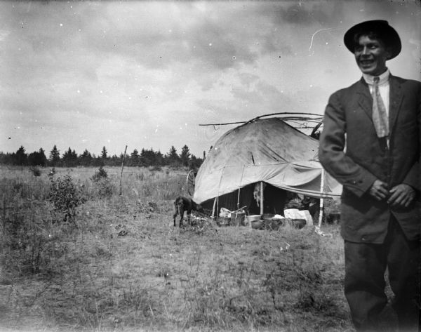 A smiling Ho-Chunk man is standing in a clearing wearing a suit, necktie, and hat. In the background is a summer Ho-Chunk lodge in a field with a dog standing nearby.