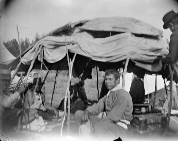 Ho-Chunk men and women sitting in and around a temporary Ho-Chunk lodge with its sides raised. Identified as the Green Cloud tent at Powwow. The Ho-Chunk man sitting on the left with his right arm in a sling is Green Cloud, and the man in center is Green Crow.