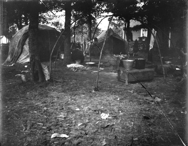 A Ho-Chunk winter lodge frame without skins, cattail matting, or fabric. In the background among trees is another Ho-Chunk lodge, tent, and wooden building. There are a few Ho-Chunk individuals nearby.