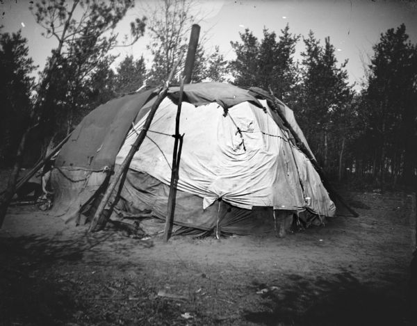 A Ho-Chunk winter lodge, with a stove pipe in the roof, in a clearing in front of trees.