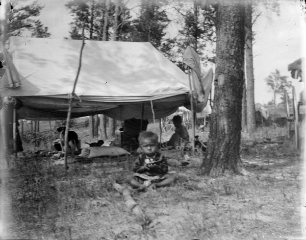 A small Ho-Chunk boy is sitting on the ground. Behind him is a Ho-Chunk summer lodge placed among trees. Inside the lodge are two dogs and another Ho-Chunk boy. On the far right is a wagon.