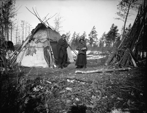 A Ho-Chunk man and woman posing standing in front of a Ho-Chunk winter lodge. The man is reportedly blind. In the background there is a horse and snow on the ground.