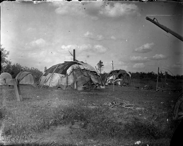 Ho-Chunk summer lodges in a field. The lodge in the center has a stove pipe coming out the top. On the left is what appears to be a grinding wheel mounted on a wooden stand. On the right are wood poles standing upright around what is probably a cooking fire.