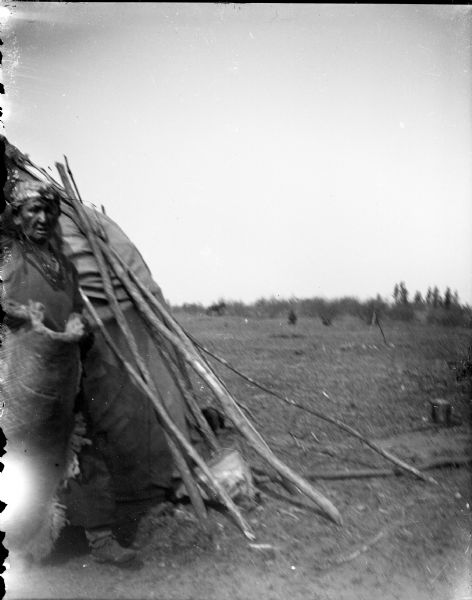 An Elderly Ho-Chunk man is posing standing in the doorway of a lodge. He has a fringed shawl wrapped around him and is wearing a head covering, necklaces, and moccasins. An open field is in the background.