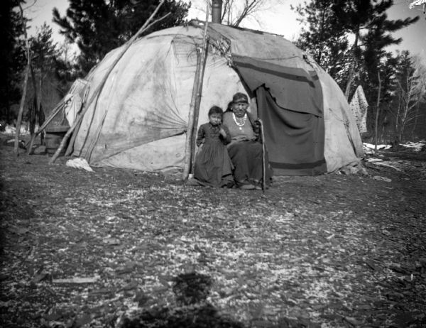 An elderly Ho-Chunk woman posing sitting and holding a cane, and a Ho-Chunk girl standing next to her, are outside the closed doorway of a Ho-Chunk lodge in front of trees. The shadow of a man, perhaps the photographer, is visible on the grass in the foreground.