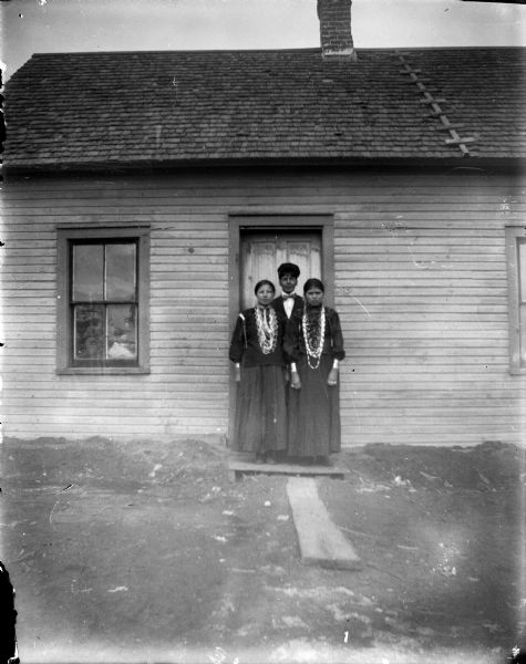 A Ho-Chunk man and two Ho-Chunk women are standing in front of the entrance door of a wooden building, probably the Mission. The man is wearing a suit, bow tie, and hat, while the women are wearing blouses, skirts, and many necklaces, file bracelets, and other jewelry. Names (l to r) Nellie Windblow, Blackdeer-Garvin, Whiterabbit-Decorah, George Bud Blackdeer-Garvin, and Nellie Windblow's sister.