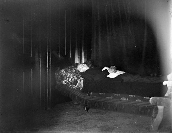 European American man layed out on a lounging sofa, dressed in a suit. Probably a corpse lying in state.