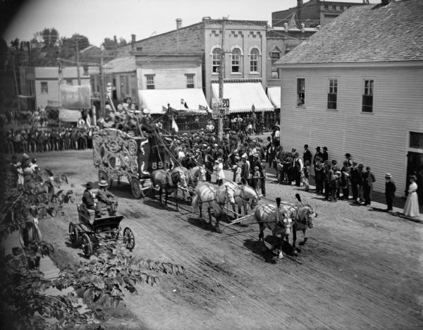 Elevated view of crowd on streets and sidewalks near an intersection watching a circus wagon pulled by a team of six horses in a circus parade in town.