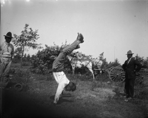 Ho-Chunk man performing a handstand in front of two Ho-Chunk men standing nearby. There is a horse-drawn wagon near trees and bushes in the background.