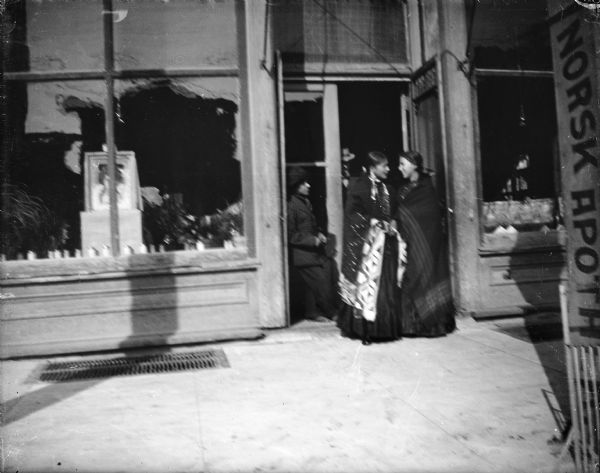 View from street of two Ho-Chunk women and a Ho-Chunk man standing in the doorway of the Werner Drugstore on the north side of Main Street between First and Water Streets.