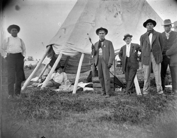 Three Ho-Chunk men and two European American men posing standing outside a teepee. The side of the teepee is raised and shows a Ho-Chunk man and woman sitting inside.