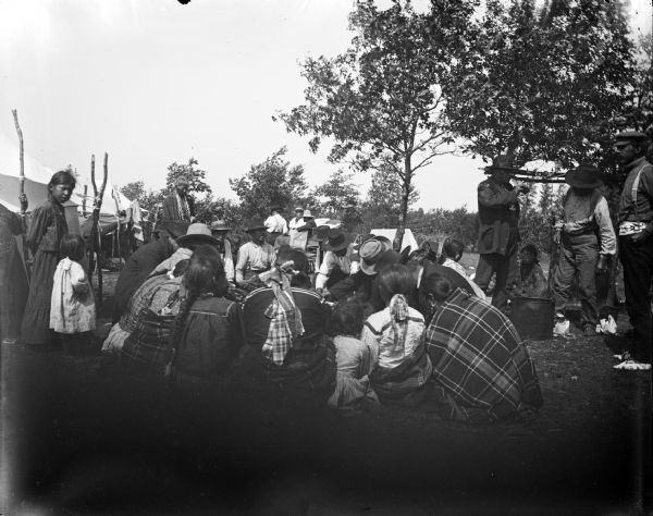 Group of Ho-Chunk men, women, and children in a circle, and in smaller groups outside the circle. They are several summer lodges. Probably a meal.