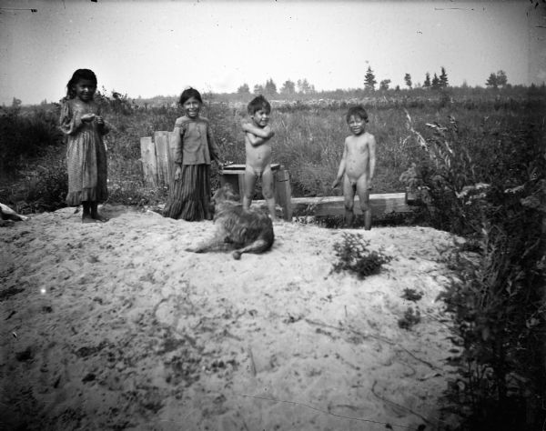Standing together in a row are two Ho-Chunk girls wearing print dresses, and two Ho-Chunk boys who are nude on a sandy pile in front of a marsh. In front of them is a dog scratching itself. The boys probably had been bathing in the marsh.