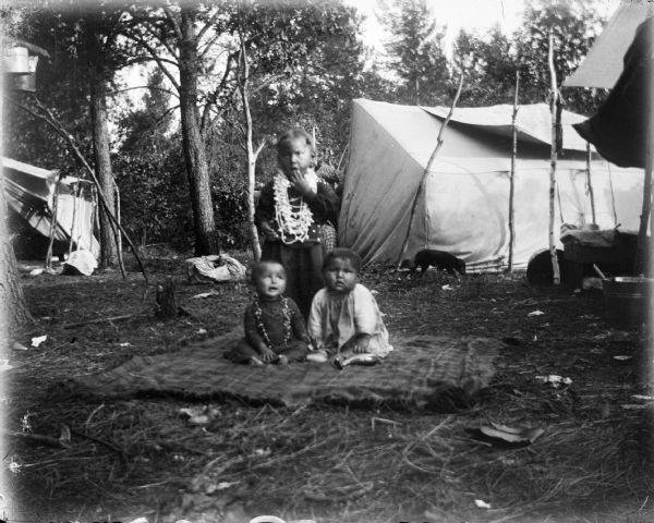 Two small Ho-Chunk children are sitting on a blanket, with a young girl wearing a long dress and necklaces standing just behind them. Canvas tents and a dog are in the background among trees.
