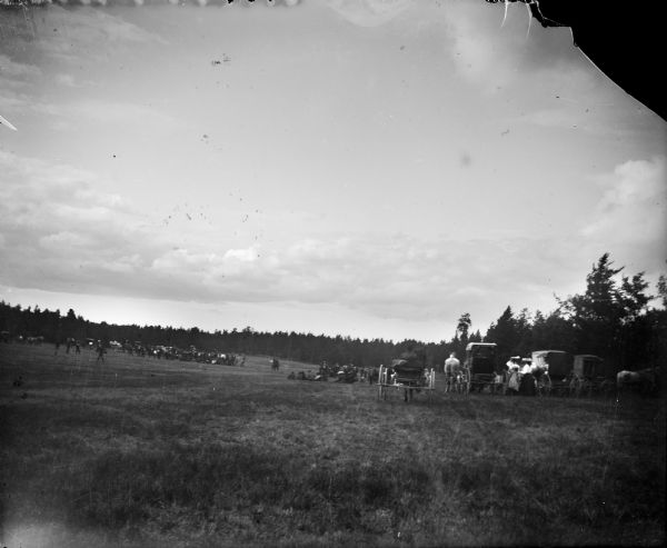View from distance of two groups of people in a open field surrounded by trees. On the right are what appears to be European Americans, with four buggies pulled by horses. Two women wearing large hats stand with a young girl in between the buggies. A group of men are sitting on the ground in the center, and in the far distance on the left are Ho-Chunk, probably gathering cranberries in a field.