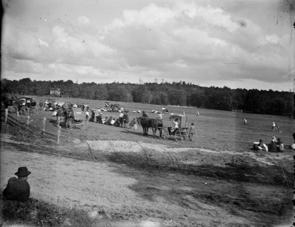 View from hill across dirt road and fence of a crowd of European Americans, and possibly some Ho-Chunk spectators. Horses are pulling buggies near the fence. The crowd is gathered in a field, where it appears men are playing baseball game.