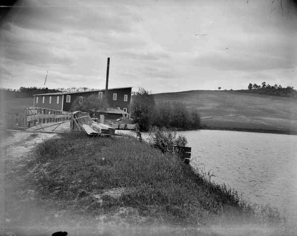View across wooden bridge to building identified as the Hixton Mill and Starch Factory on the edge of a body of water, probably a river. In the background is a hill with trees on top.