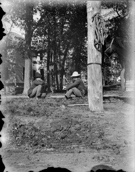 Two men, possibly Ho-Chunk, wearing hats are sitting on the ground near a board sidewalk underneath some trees. A building is behind them on the left, and on the right a horse is tied by it's harness to a power line pole, with a portion of possibly a street sign above a hook holding a coil of rope.