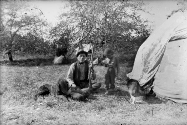 Portrait of a Ho-Chunk man sitting cross-legged on the ground outside of a tent, holding a gun and wearing a vest and hat. A dog is on the ground near his right knee, with what appear to be two or three puppies. On his right is a Ho-Chunk boy standing next to him, who is wearing a hat and holding something in his hand. Near the tent doorway there is another dog. In the background near some trees is a blurred object, possibly another person.
