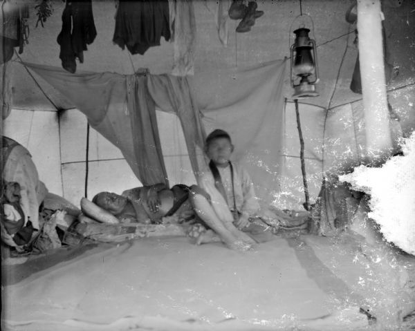 An elderly Ho-Chunk man is laying down and sleeping behind a Ho-Chunk boy who is sitting, possibly inside of a Ho-Chunk lodge or tent. A lantern is hanging from the ceiling along with some clothing and a pair of shoes.