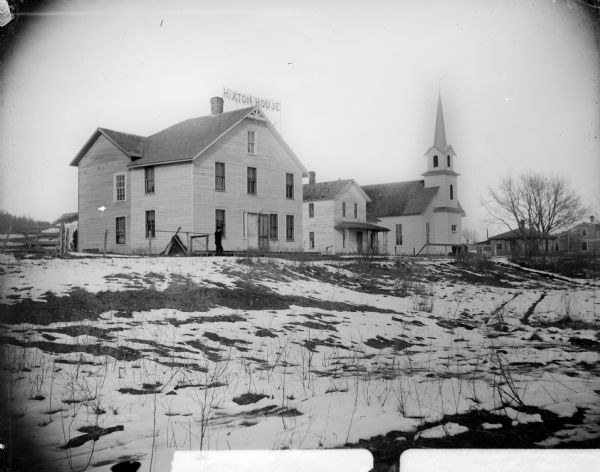 Portrait of several buildings and a snowy landscape, probably in Hixton. A man is standing outside the first building on the left, which has a sign on the roof that reads: "Hixton House," and is probably a hotel built by Charlie Northrup about 1912. Next from the left is a house with a porch, and to the right of that is a Methodist Church that later became a Presbyterian Church. Other buildings are on the right after the church.