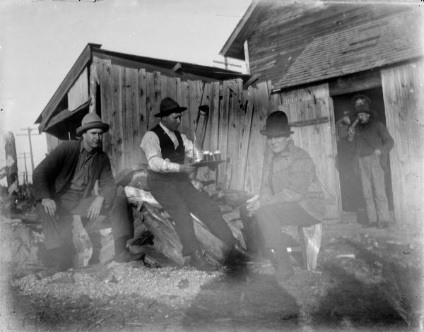 Three men are posing sitting on a woodpile near some wooden buildings. The man in the center is wearing a vest and hat and is holding a platter with a glass bottle and several glasses filled with a beverage. The other two men on his right and left are wearing suit coats and hats. In the background in the doorway of a building three other men are standing. In the foreground is the shadow of a man, perhaps the photographer.