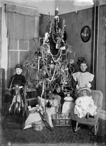 Portrait of a boy and girl posed by a Christmas tree. The boy is sitting on a toy horse on the left and is wearing a military style hat and holding a toy gun. The girl is poing on the right standing behind a doll on a chair. She is wearing a large bow in her hair and a dress. The tree is decorated with ornaments, strings of popcorn, and tinsel. Underneath the tree are various toys including a tea set, a broom, dolls, a drum, a miniature stove with pots and pans, and a toy gun.