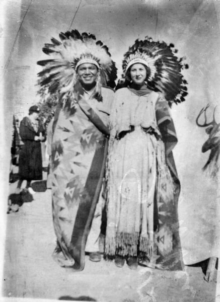 Copy print portrait of a Ho-Chunk man, standing on the left next to a European American woman near a tent. Both of them are wearing Ho-Chunk regalia including large feather headdresses. Another European American woman is standing in the background.