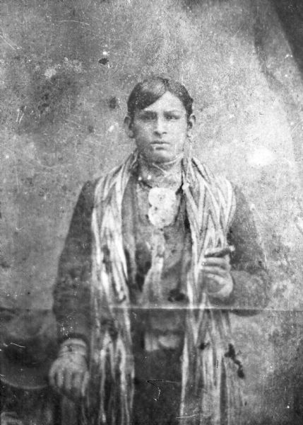 Three-quarter length copy photograph of a Ho-Chunk man posing standing. He is wearing full regalia, including a finger braided sash, and holding a calumet pipe. George Funmaker (wox 'Ai Le k, Bear clan) and the sash is still reportedly in the family.