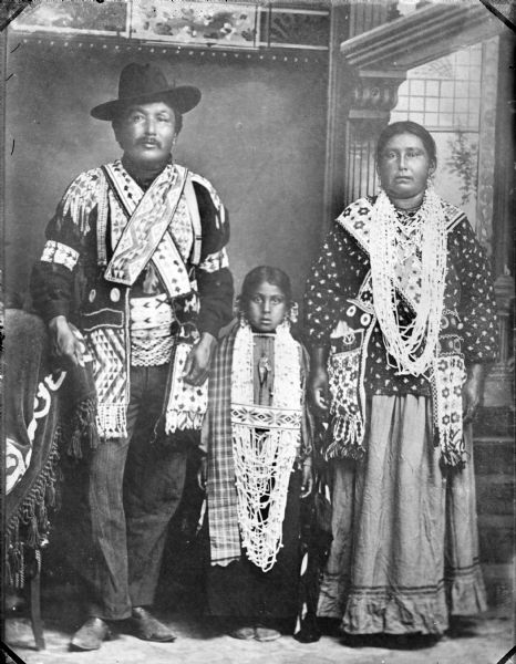 Studio portrait of a Sioux man, Ho-Chunk woman, and Ho-Chunk girl, all posing standing in front of a painted backdrop and wearing full regalia. Identified as David Goodvillage and his wife, he was a Sioux and she was the daughter of Chief Blackhawk, their grandchild was Mabel White.