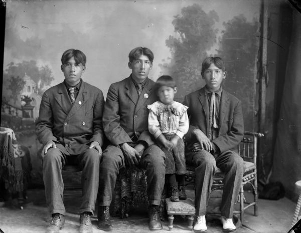 Studio portrait of three young Ho-Chunk men posed sitting in front of a painted backdrop and wearing suits, one holding a small boy. The male children of the Lowe family, from left to right: Theodore Lowe, Albert Lowe, and Sam Lowe, the child is Gilbert Lowe.