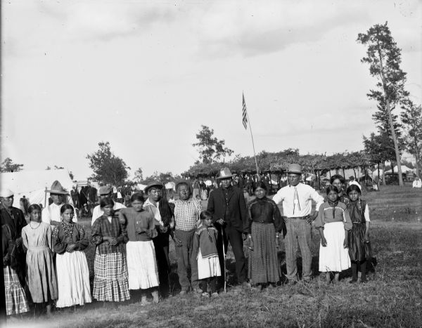A group of Ho-Chunk men, women, and children posed standing in a field. In the background behind them is the front of an arbor with a United States flag, and tents surrounding it. Possibly a powwow near Decorah Cemetery. Identified, left to right as: unidentified, Mrs. Garvin (behind), unidentified, Emma Funmaker (behind), unidentified, unidentified, unidentified, unidentified, Joe Bearheart (child), unidentified, unidentified, Lucy Lowe Yellow Bank, unidentified, Annie Winneshiek (behind), unidentified, and unidentified.